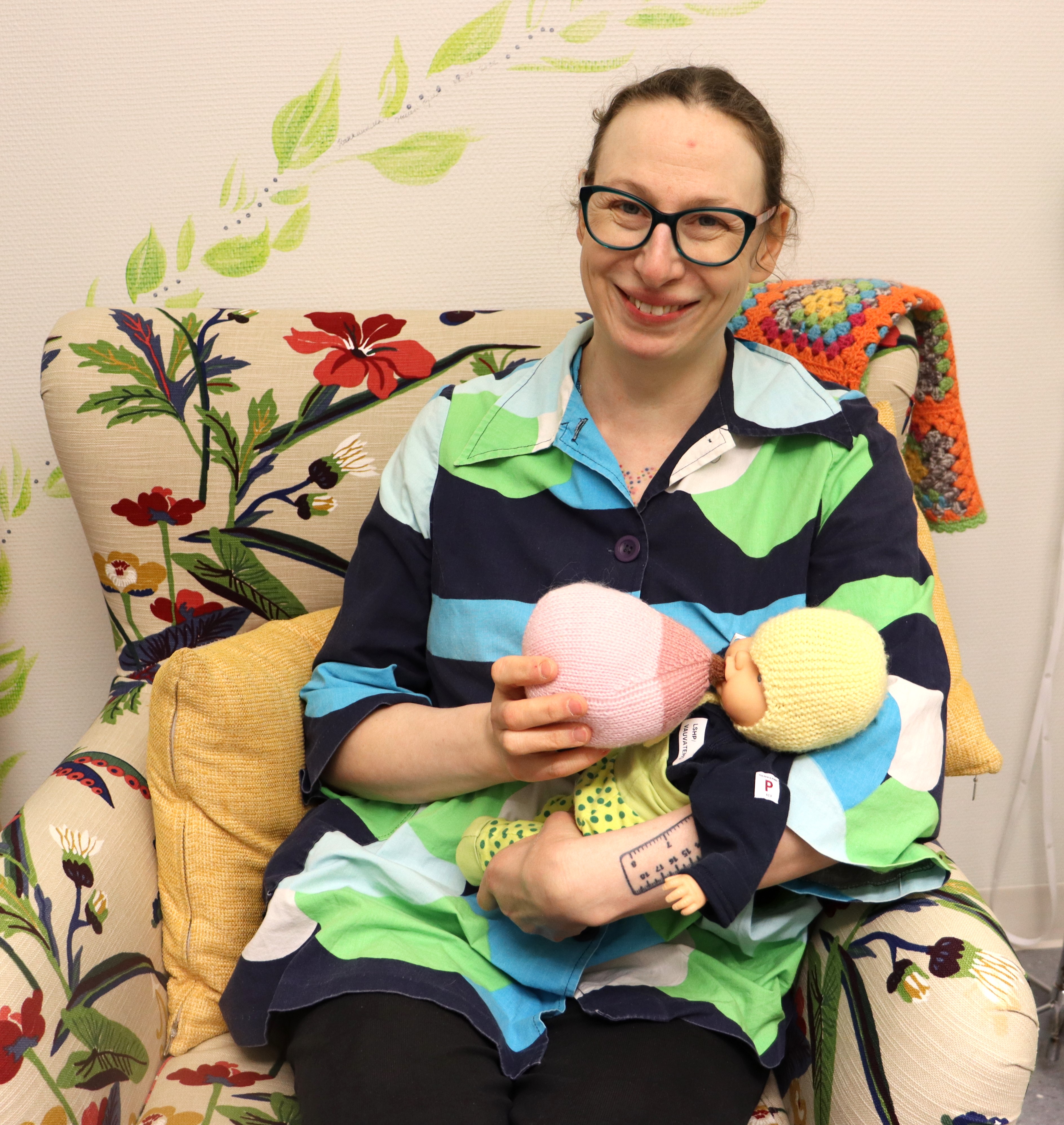 Kätilö Hanna Ekblom istuu nojatuolissa käsissään vauvanukke ja neulottu opetusnänni. 