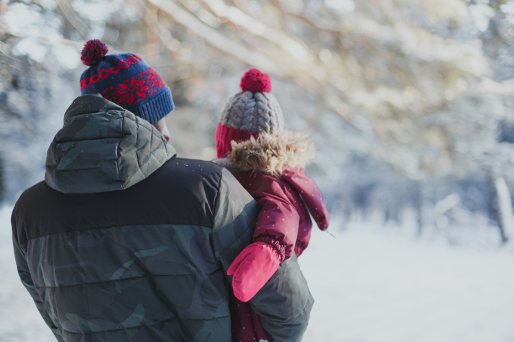 Talvivaatteisiin pukeutunut isä pitelee sylissään punaiseen haalariin pukeutunutta pientä lasta. Isä ja lapsi ovat selin kuvaajaan päin ja katselevat talvista maisemaa.