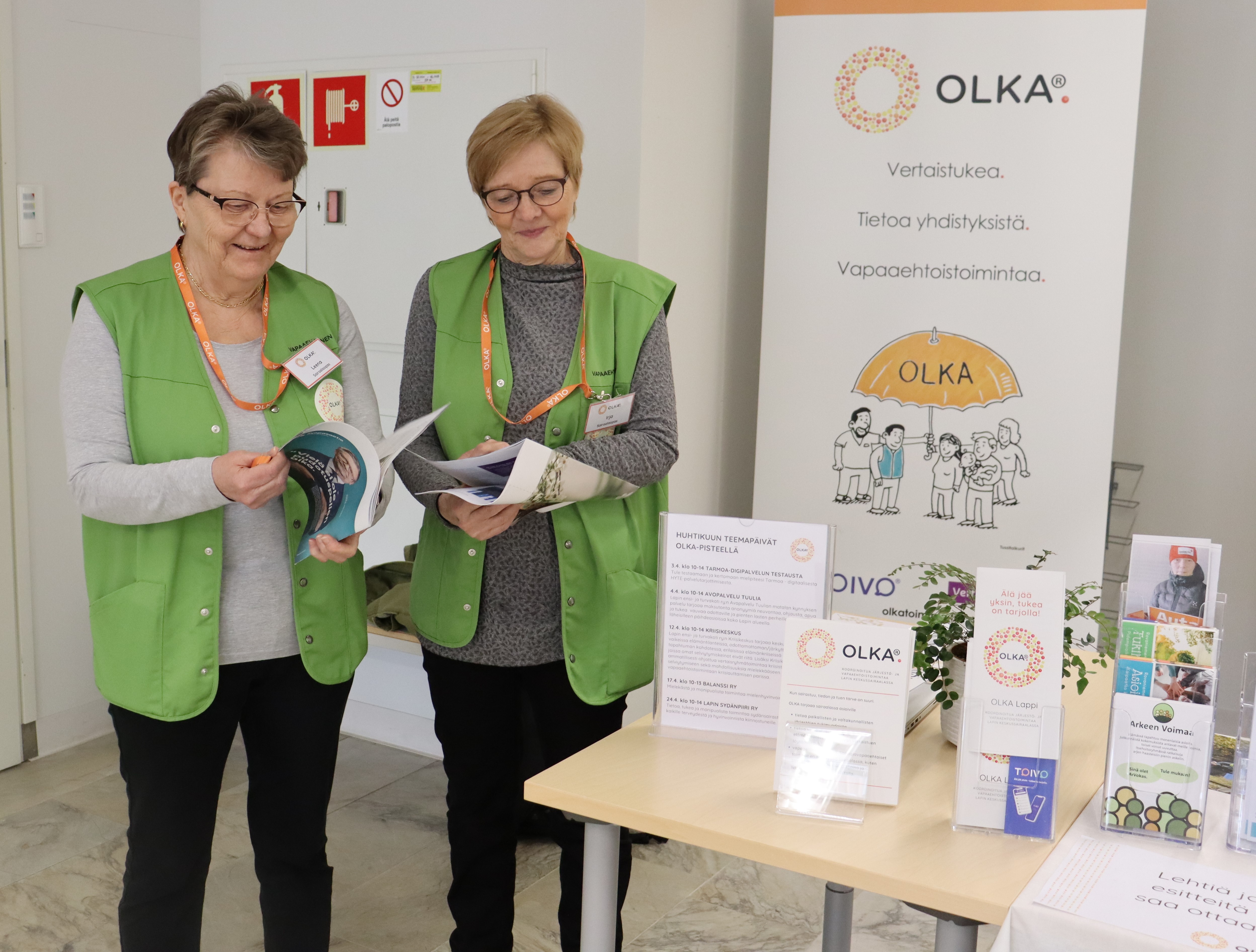 Kaksi vihreisiin liiveihin pukeutunutta naista lukee lehtiä Lapin keskussairaalan aulassa. Taustalla OLKA-pisteen roll up, jossa kerrotaan vapaaehtois- ja järjestötyöstä.