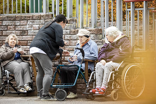 Kolme vanhusta istuu ulkona pyörätuoleissa. Hoitaja tarjoaa yhdelle vanhukselle kahvikuppia.