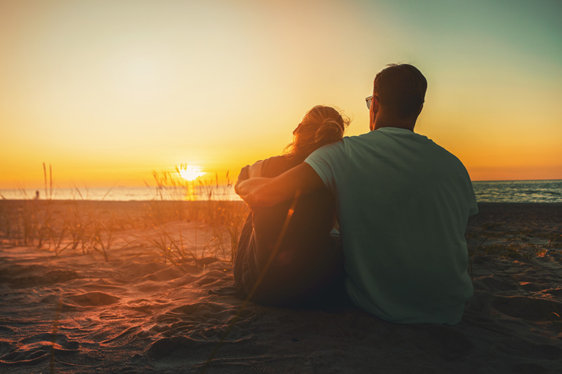 Kaksi ihmistä rannalla istumassa auringonlaskussa.